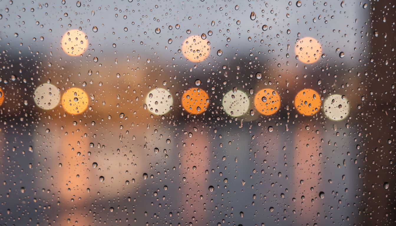 Rain drops on glass with blurred warm city lights behind — the ASMR rain-on-window aesthetic